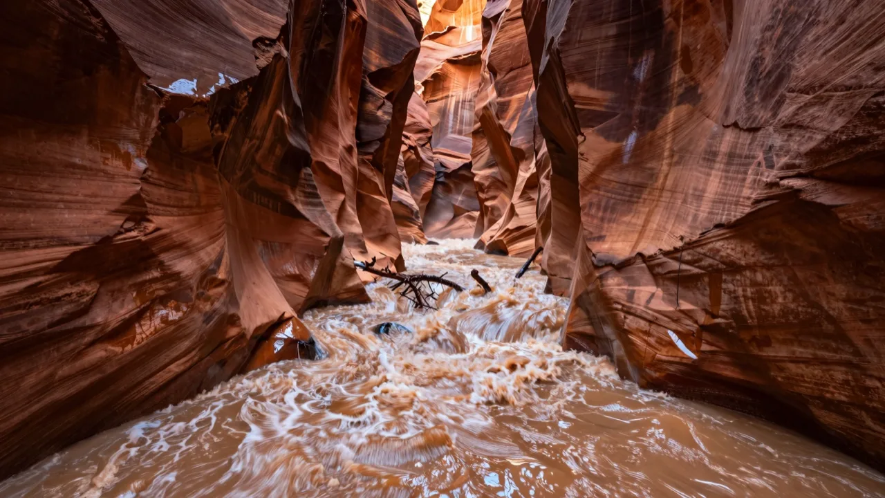 Muddy floodwater surging through a narrow red sandstone slot canyon under storm light